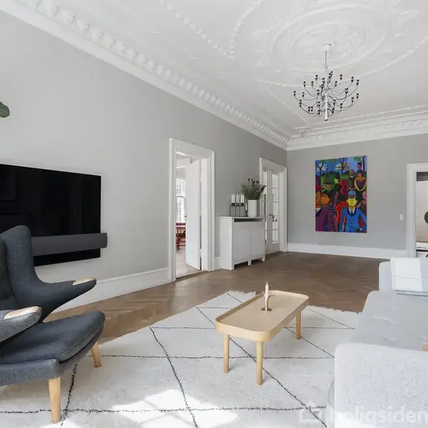 Stue med grå vægge, en grå lænestol ved siden af et TV på væggen, en lys sofa, et sofabord i træ, kunst på væggen, og en stor grøn plante.