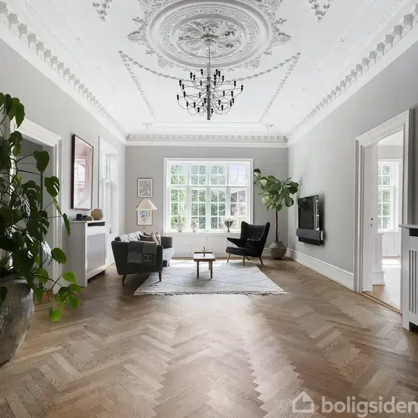 Stue med hvid sofa og sort lænestol centreret på et tæppe, under en sort lysekrone. Store vinduer i baggrunden slipper lys ind. Grønne planter skaber en frisk atmosfære.