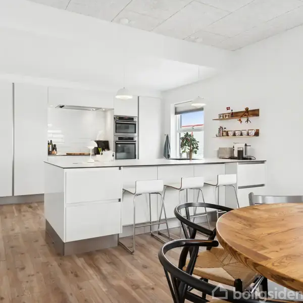 Køkken med en hvid køkkenø og barstole beliggende på trægulv. Rundt bord med stole i forgrunden, planter på vindueskarmen og hylder med køkkenredskaber skaber en lys og elegant atmosfære.