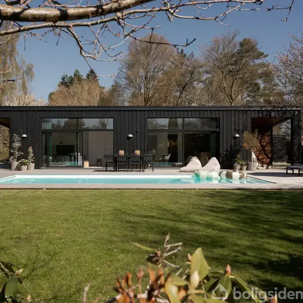Moderne sort hus med store vinduer, stående ved en swimmingpool. Liggestole og parasol arrangeret ved poolen i en have omkranset af træer og buske.