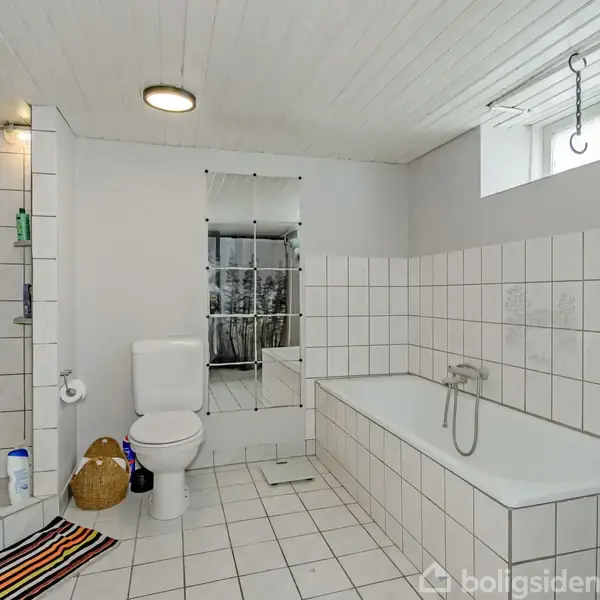 Toilet og badekar med hvidt flisegulv og -vægge, placeret i et rummeligt badeværelse. Spejl og vindue i baggrunden. Håndklæder hænger ved badekarret, og kurv ved toilettet.