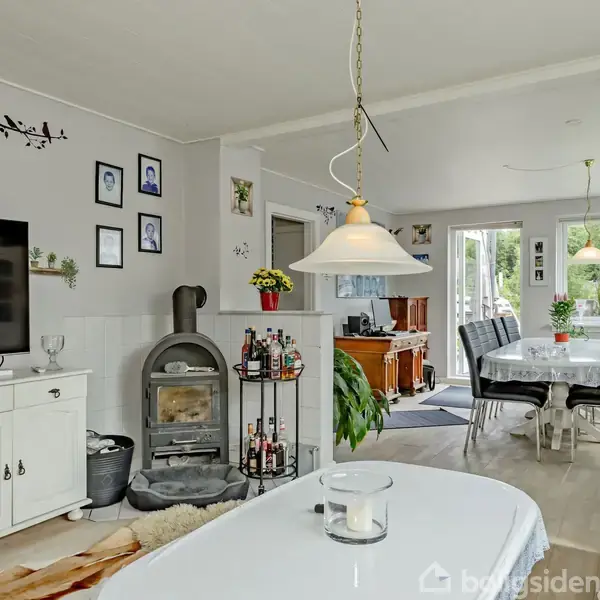 En stue med en brændeovn og fjernsyn i midten. Et spisebord står foran store vinduer med udsigt til en have. Flere planter og billeder dekorerer rummet.