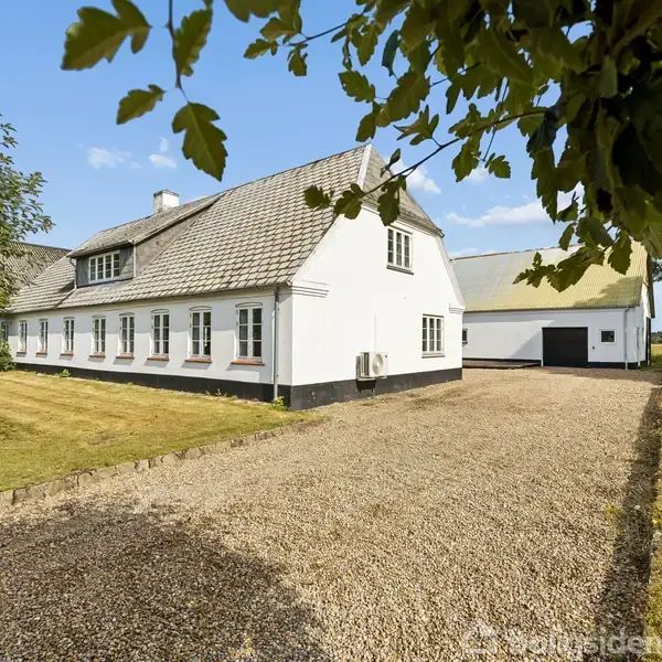 En hvidmuret landejendom med grått tag står for enden af en grusvej, omgivet af træer og græsplæner under en klar, blå himmel.