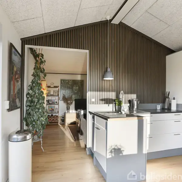 Køkken med hvidt skab og grå bordplade, der står stille. Lampe hænger over bordet. Plante ved siden af åbningen til en stue med billeder på væggene.