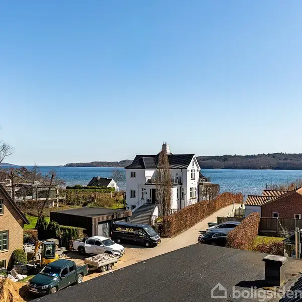 Huse står stille i et boligkvarter med havudsigt i baggrunden. Flere biler er parkeret langs vejene blandt bygningerne. Atmosfæren er rolig og kystnær.