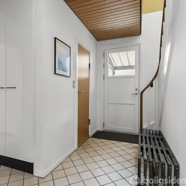 Entré med flisegulv og træloft. En hvid dør med glas fører udenfor. Til venstre er skabe og en åben dør til et værelse. Trappe med gelænder til højre.