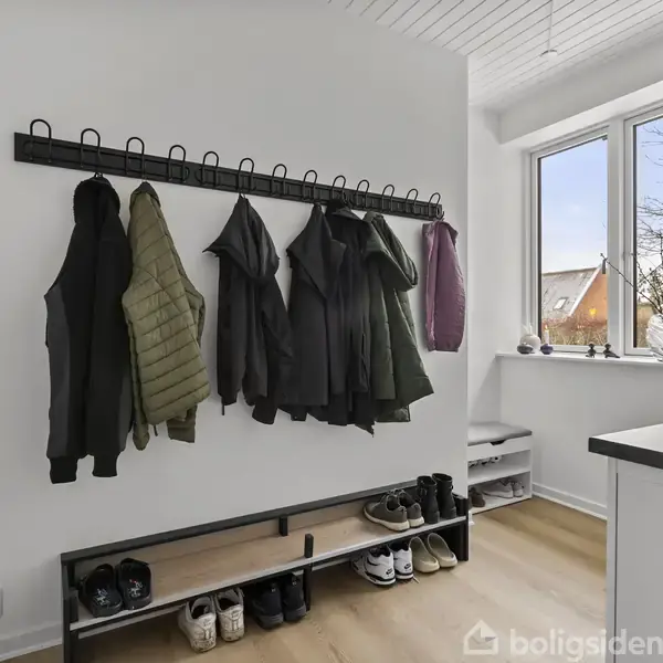 Jakker hænger på en knagerække, flere par sko er placeret på en hylde under dem. Den lyse entré har et stort vindue med udsigt til hustage og træer.