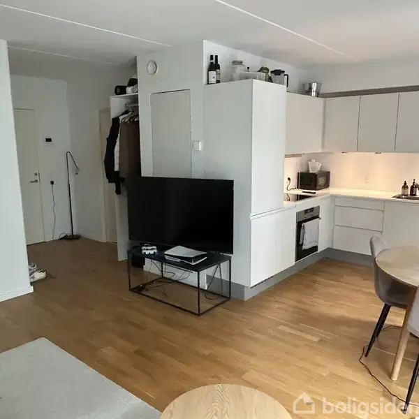 Fladskærms-TV står på et stativ i hjørnet af en stue, foran en moderne åben køkken med hvide skabe. Tæppe og spisebord med stole er placeret på trægulv.