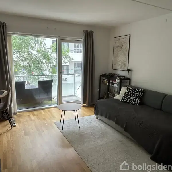 En stue med en grå sofa til højre, et lille rundt bord foran og en computer ved et skrivebord til venstre. Store glasdøre fører ud til en altan med træer udenfor. Ingen tekst fundet.