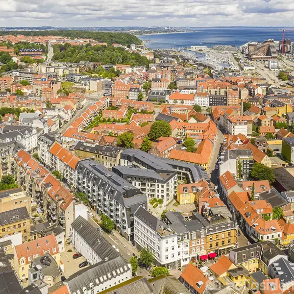 Byområde med røde tegltage og bygninger ses fra oven, omgivet af grønne områder og en havn i baggrunden under en skyet himmel.