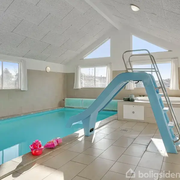 Indendørs swimmingpool med en rutsjebane fører ned i vandet. Legetøj ligger ved kanten. Rummet har store vinduer, beige vægge, og lyse fliser. Loftet hælder opad og har loftslys.