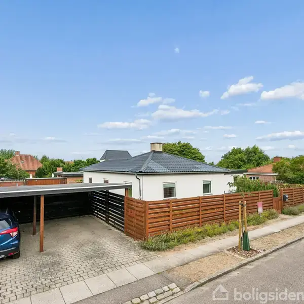 En hvid villa med mørkt tag ses bag en carport, hvor en blå bil holder parkeret. Området er roligt med grønne hække og flere boliger i baggrunden.