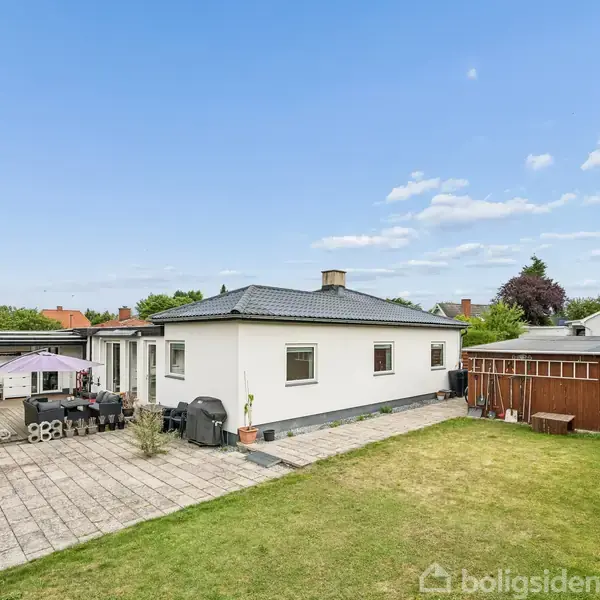En hvid bygning med fladt tag står på en grøn græsplæne. En flisebelagt terrasse med havemøbler og grill omkranser huset. Træskur til højre, omgivet af beplantning.