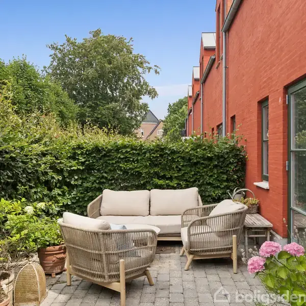 Terrassemøbler med sofagruppe og stole står på flisebelagt udeområde. Placeret nær rød murstensbygning omgivet af frodig hæk og blomster. Solskinsdag skaber lys og skyggeeffekter.