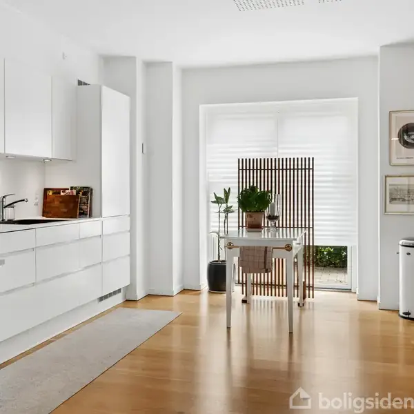 Et moderne køkken med hvide skabe og en bordplade med brødrister og skærebræt; træbord i midten med planter; store vinduer danner lys baggrund.
