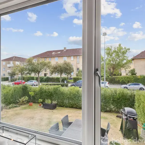 Vindue rammer udsigt over en have med terrasse og grill; i baggrunden ses en vej, høje hække og bygninger med røde tage. Biler kører langs vejen.
