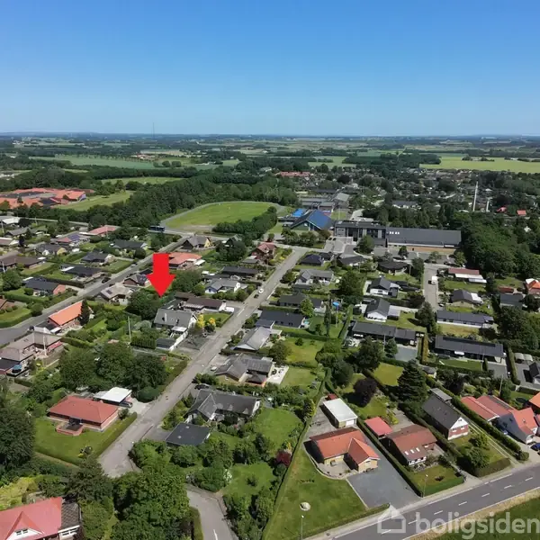Et boligområde ses fra luften, hvor en rød pil peger på et bestemt hus blandt mange. Området er omgivet af grønne marker under en blå himmel.