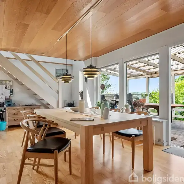 Spisebord med stole står i lys stue. To lamper hænger over bordet. Store vinduer og dør fører til terrasse med udsigt over grønne omgivelser. Trappe leder opad i baggrunden.