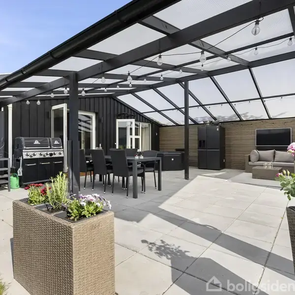 Terrasse med spisebord og otte stole under en overdækning i glas. Der er en grill til venstre og en lounge-sofa i baggrunden. Store plantekasser definerer området.