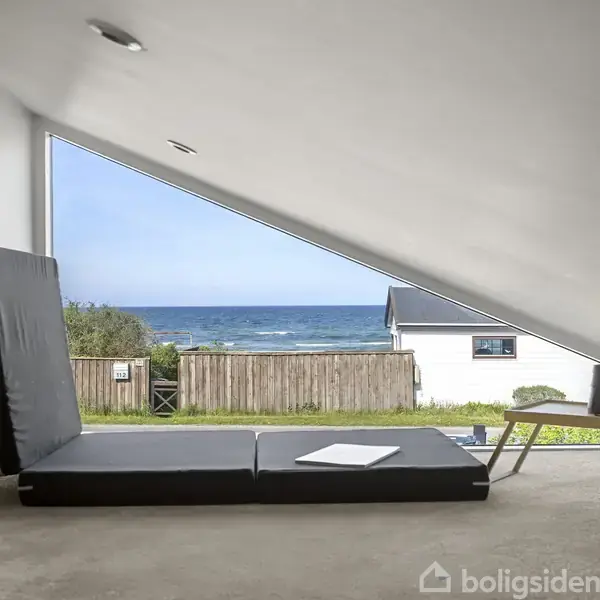 Madras placeres tæt på stort skråvindue med udsigt til havet; udenfor ses græs, hegn og et hvidt hus.