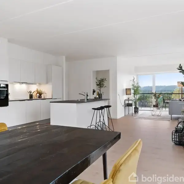 Køkkenø med sorte barstole står i centrum af et moderne, lyst køkken. I baggrunden er der en stue med åben altandør, der giver udsigt til en balkon med grønne planter.