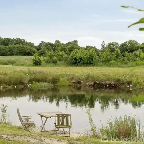 Et bord og to stole står stille ved bredden af en lille sø. Omkring dem ses græs og skovbevoksning under en klar blå himmel.