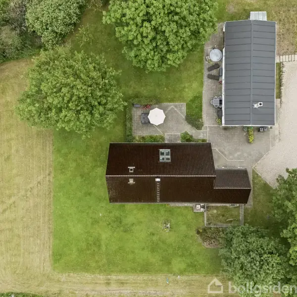 Hus med mørkt tag står i grøn have omkranset af træer og græsplæne. Ved siden af ses en terrasse med hvid parasol. Grusvej fører til anden bygning med gråt tag.