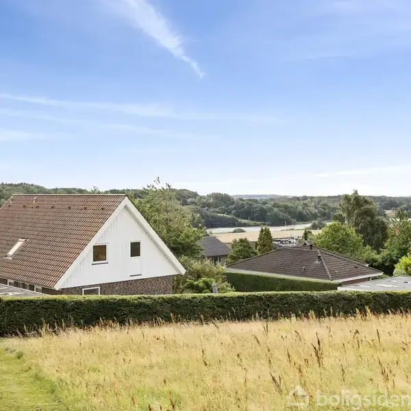 Et hus med rødt tag står stille omkranset af en grøn hæk og græsplæne. I baggrunden ses et landskab med skove og bakker under en klar blå himmel.