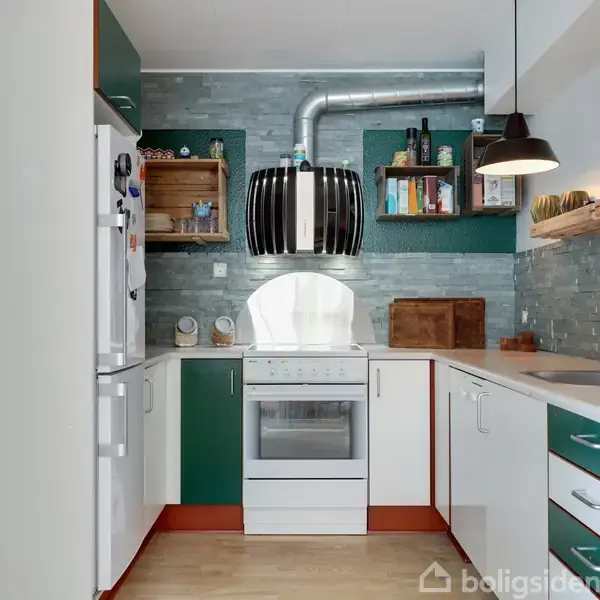 Komfur med moderne emhætte, omgivet af grønne og hvide skabe. Skærebræt og bøger på hylderne over køkkenbordet. Loftlampe hængende ned over stålvaske i et lyst, indbydende køkken.