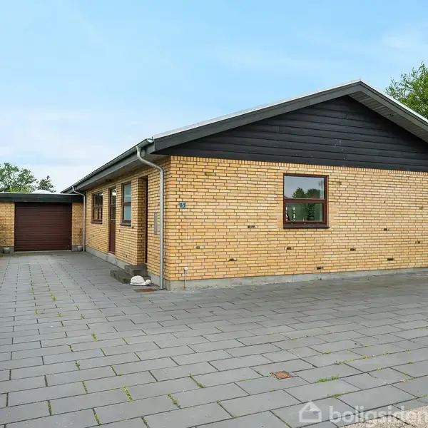 En gulstensbungalow med tilhørende garage står stille. Omkringliggende flisebelægning og grønne hække omkranser huset. Himlen er klar og blå.
