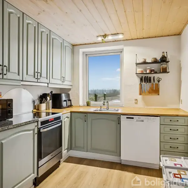 Et køkken med lysegrønne skabe og træbordplade. En ovn og mikroovn til venstre, vindue med udsigt midtfor, og hylder med knive og krydderier til højre.