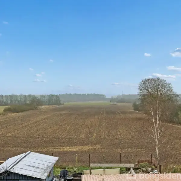 En åbent mark strækker sig mod horisonten under en klar blå himmel med spredte skyer; i forgrunden ses en del af et metal- og trætag fra en bygning.