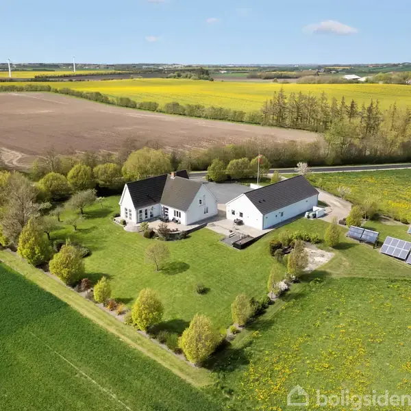 En hvid landejendom med sort tag står midt i en grøn have med solpaneler. Baggrunden viser marker og gule blomster, indrammet af en landlig, åben natur.