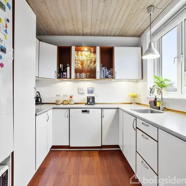 Køkken med hvide skabe. Køkkenredskaber og planter på bordpladen, vindue til højre. På køleskabet ses magneter og noter. Trægulv og lyst træloft.