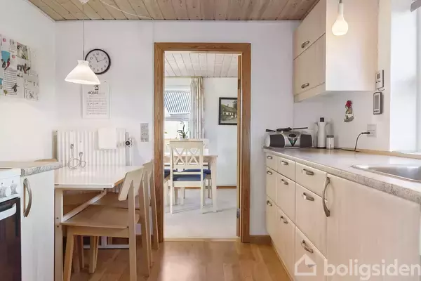 Køkken med lyse træmøbler, indbygget komfur, og opvaskebord; en åben dør fører til en spisestue med hvidt spisebord og stole. Vægge dekoreret med ur og billeder.