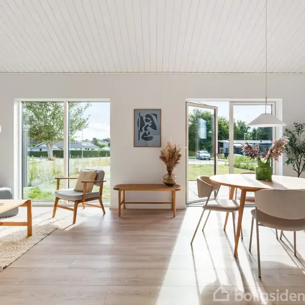 En lys stue med moderne møblering. Et rundbord med stole står nær en åben dør mod en have. Træmøbler, planter og kunst på væggen skaber en varm atmosfære.