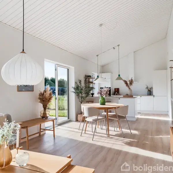 Stue med lyse træmøbler, en stor papirlampe hængende, og udsigt til haven gennem glasdøre. Et lille spisebord står foran et lyst køkken. Dekorative planter og blomster fylder rummet.