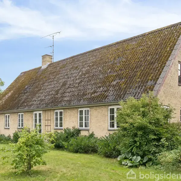 Et langstrakt hus med mosgroet sadeltag står stille i en frodig have. Vinduerne er symmetrisk placerede, og en flagstang står til venstre for huset.