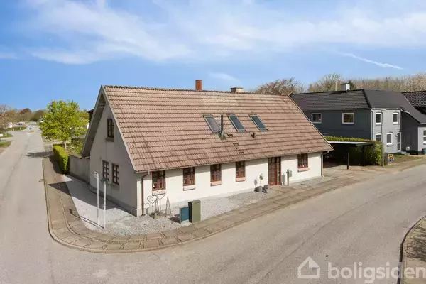 Et hvidt hus med rødt tegltag og tagvinduer står ved en vej, omgivet af et flisebelagt område og hække. Nabohusene er synlige i baggrunden under en blå himmel.