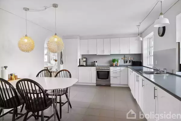 Et moderne køkken med hvide skabe, en sort bordplade og sort/hvide gulvfliser. Et rundt hvidt spisebord med fire mørke træstole står under to dekorative lamper. En døråbning fører til et andet rum.