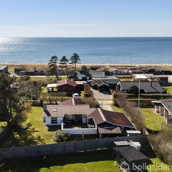 Hus ved kysten med rødbrunt tag ligger i et boligområde. Haven er omgivet af hegn. I baggrunden ses havet og stranden under en klar blå himmel.