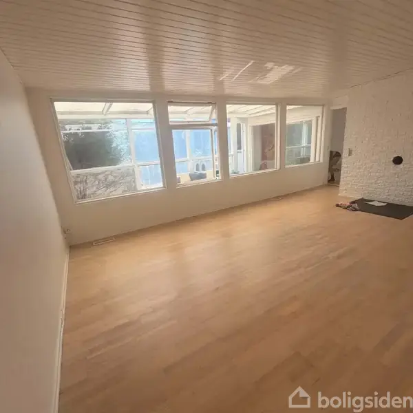 Et tomt rum med trægulv har store vinduer langs væggen, der giver udsigt til en tilstødende overdækket terrasse. Væggene er lyse, og der er en murstensdetalje til højre.