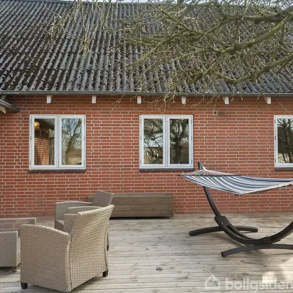 En terrasse med trædæk, hvor en hængekøje på stativ står ubrugt ved siden af kurvemøbler. Baggrunden er en rød murstensbygning med flere vinduer og et trætag.