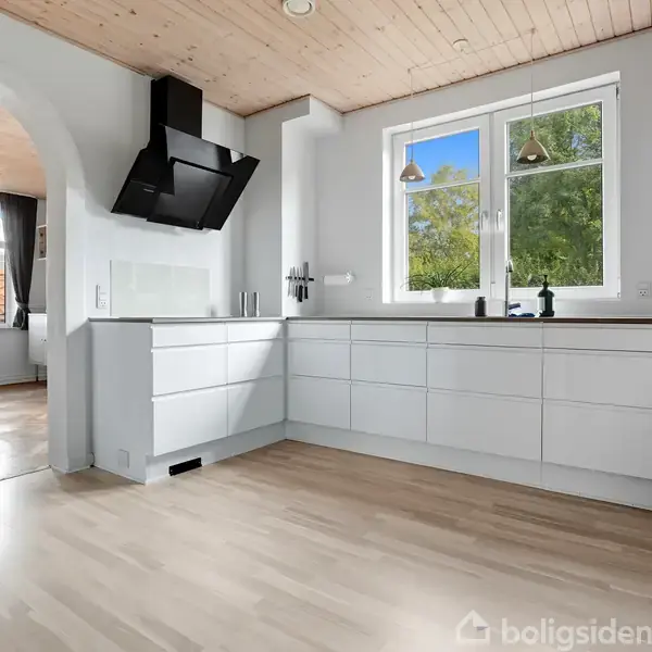 Et lyst køkken med moderne, hvide skabe og sorte håndtag. En stor emhætte hænger over kogepladen. Vindue med udsigt til grønne træer. I baggrunden ses en spisestue gennem en bue.