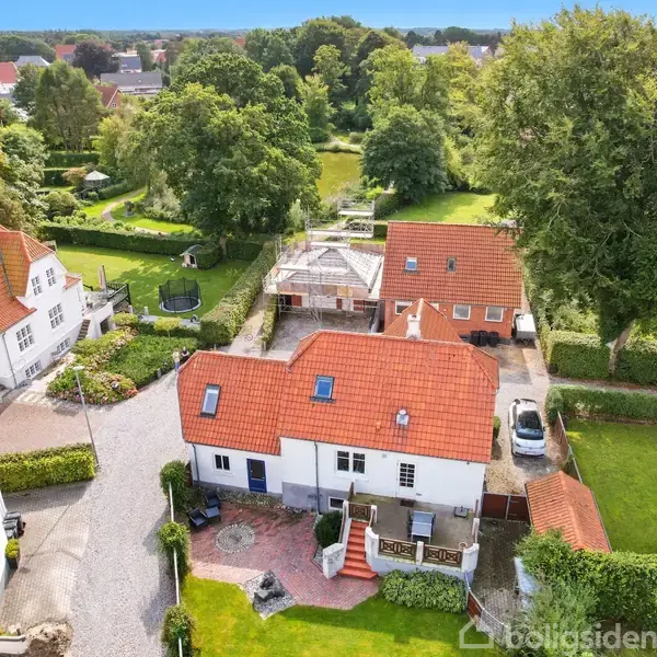 Et hvidt hus med rødt tegltag står centralt. Det omkranses af en velplejet have med grøn plæne og træer, og der er andre karakteristiske huse med røde tage i nabolaget.