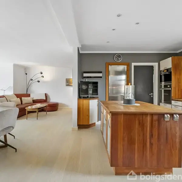 Køkkenø i træ fungerer som central arbejdsstation i et åbent opholdsområde med spisebord og lysekrone på venstre side, samt en beige sofa i baggrunden.