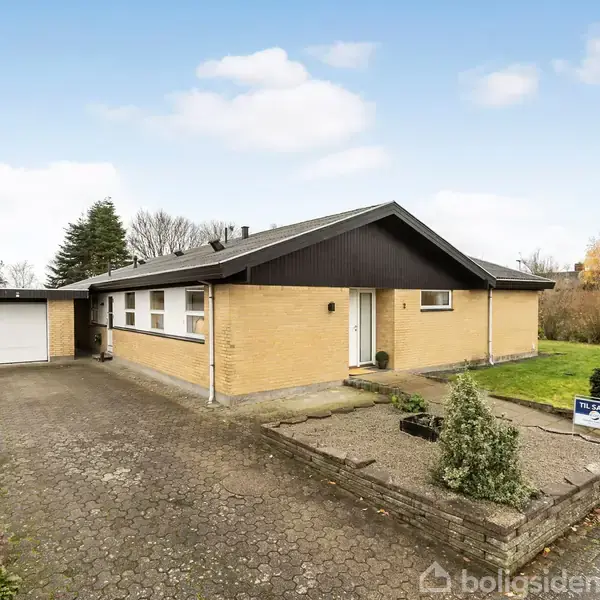 En gul murstensbungalow med sort tag står på en flisebelagt indkørsel ved siden af en garage. Foran huset er en lille have med en ejendomsmægler-skilt: "Ny-pris".