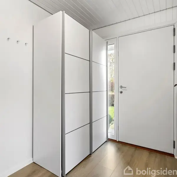 Hvid skab står stille i entre med trægulv, ved siden af en hvid dør med glasparti. En lille radiator med en plante er synlig. Der er ingen tekst.