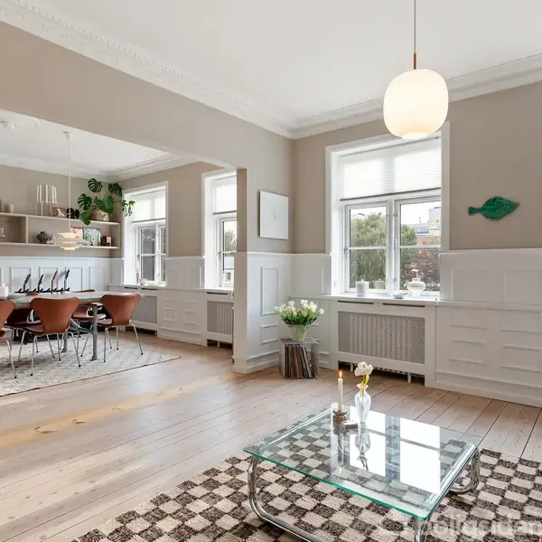 Stue med glasbord og sofa, indrettet i lyse farver. Til venstre er spisestue med træstole og hylder. Store vinduer giver naturligt lys. Blomsterdekorationer og kunst tilføjer detalje.