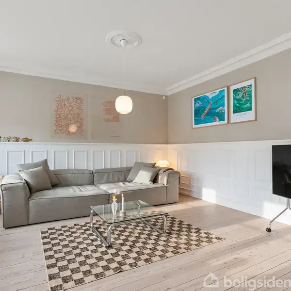 En lys stue med en grå sofa, glasbord med blomster, og en sort fladskærms-tv. To farverige billeder hænger på væggen over en hvid panelvæg.